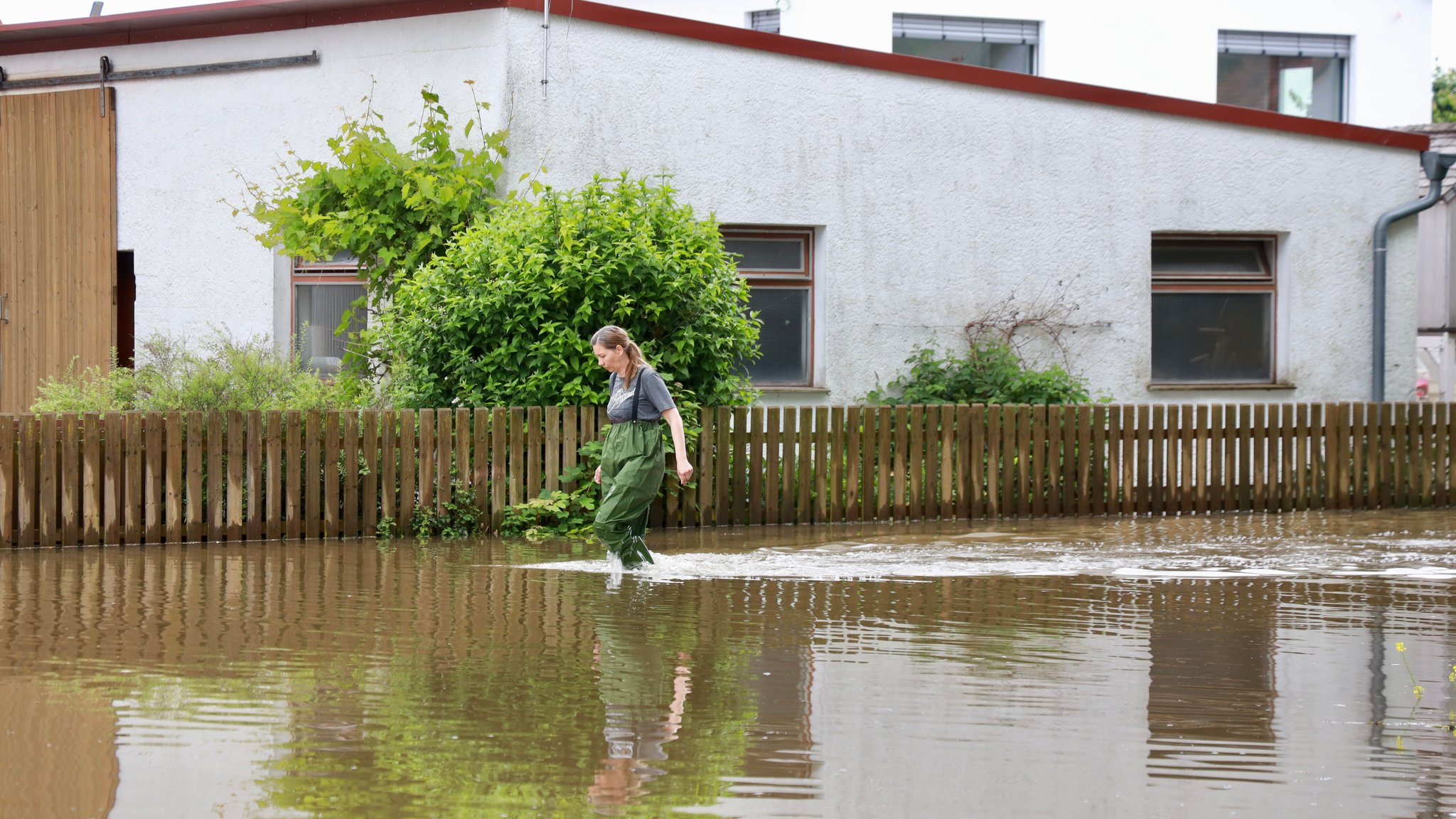 Eine Frau in wasserfesten Hosen läuft durch braunes Hochwasser, im Hintergrund ein Haus.